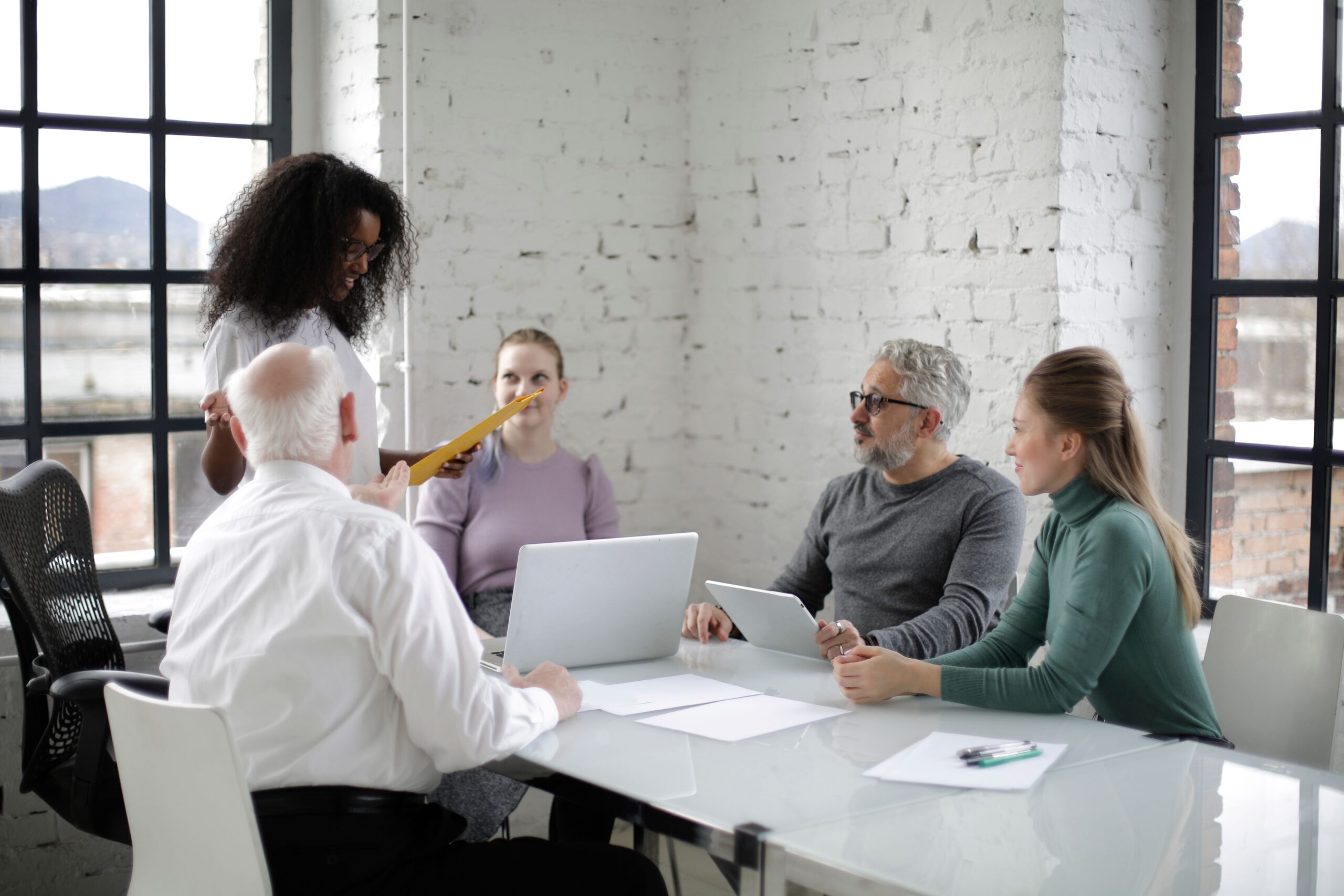 Five people having a meeting around a long table with papers, with one woman stood up speaking to the group.